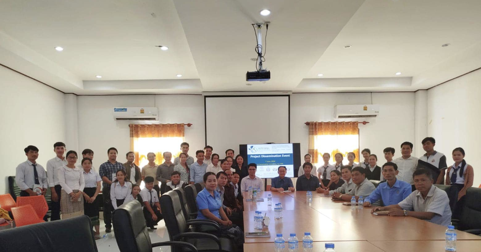 Group photo of university instructors and students in front of a slideshow projection in a meeting room