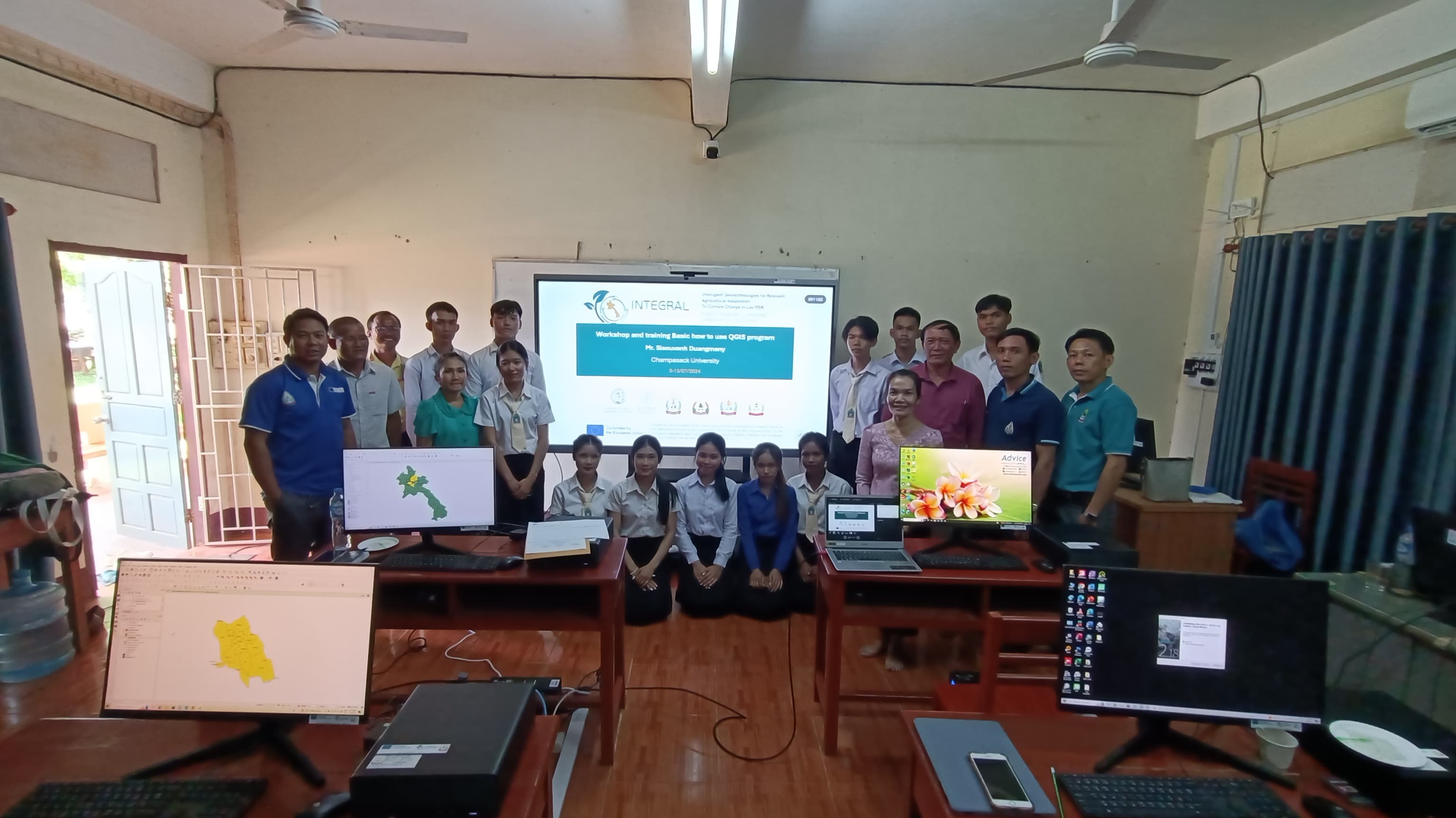 Group photo of university instructors and students in a computer lab with maps of Lao PDR on some screens